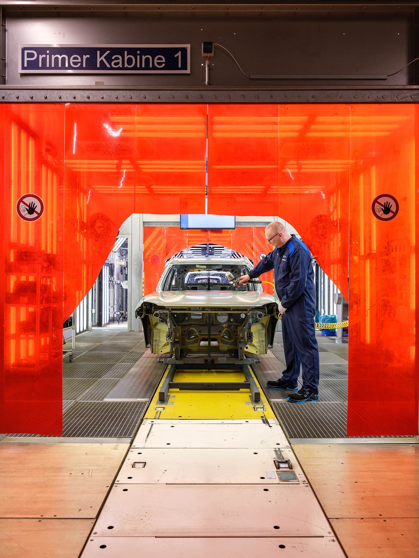 Factory worker in blue overalls stands to the side of an unfinished car body shell, framed by the neon orange PVC panel that encloses the production cabin.
